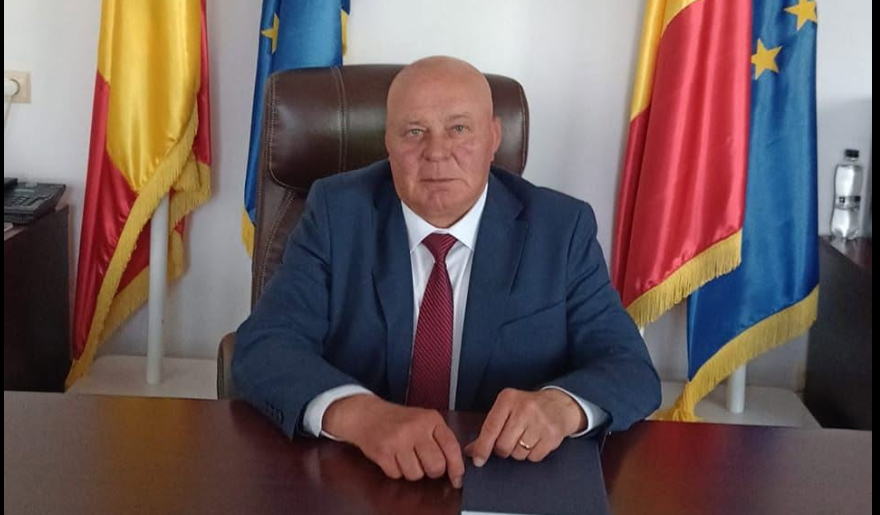 Bald man in a blue suit and red tie sits at a wooden desk in an office, with flags behind him and hands resting on the desk.