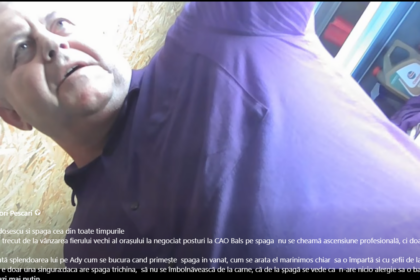 Man in a purple shirt leaning toward the camera in a workshop or shop, shelves with cleaning products visible in the background.
