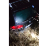 Dark hatchback crashed in tall dry grass at night, rear of the car visible with red taillights.