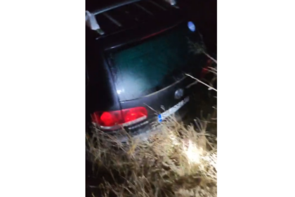 Dark hatchback crashed in tall dry grass at night, rear of the car visible with red taillights.
