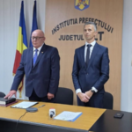 Two men in dark suits stand behind a desk in a government press briefing room, with Romanian flags in the background and a prefecture sign nearby.