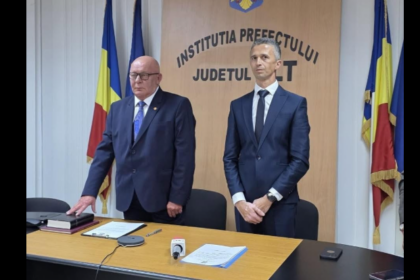 Two men in dark suits stand behind a desk in a government press briefing room, with Romanian flags in the background and a prefecture sign nearby.