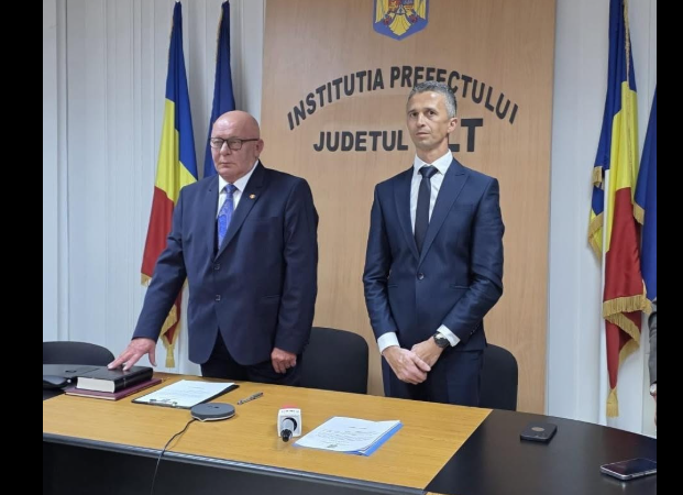 Two men in dark suits stand behind a desk in a government press briefing room, with Romanian flags in the background and a prefecture sign nearby.