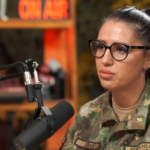 Woman in camouflage military uniform wearing glasses speaks into a studio microphone with an 'ON AIR' sign in the background.
