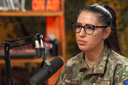 Woman in camouflage military uniform wearing glasses speaks into a studio microphone with an 'ON AIR' sign in the background.