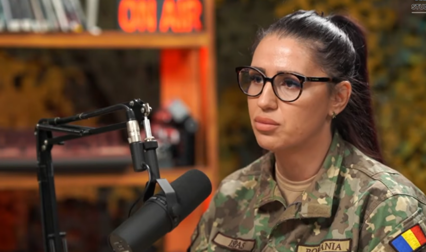 Woman in camouflage military uniform wearing glasses speaks into a studio microphone with an 'ON AIR' sign in the background.
