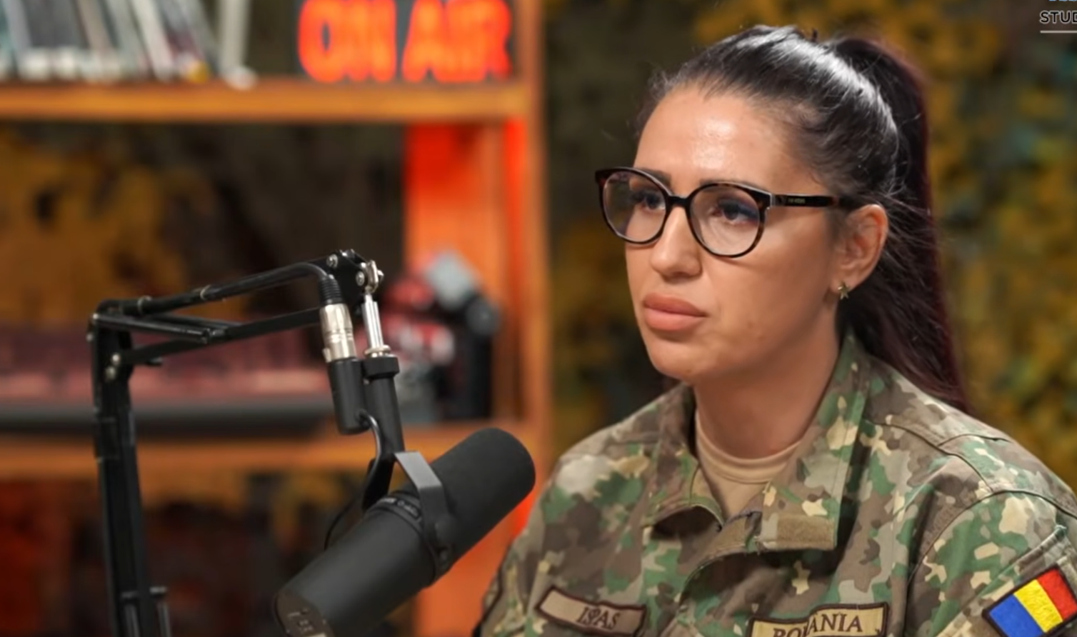 Woman in camouflage military uniform wearing glasses speaks into a studio microphone with an 'ON AIR' sign in the background.