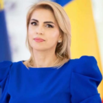 Professional woman in a blue dress gazing slightly to the left, with blurred flags in the background indicating a formal event or press setting.