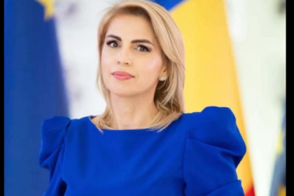 Professional woman in a blue dress gazing slightly to the left, with blurred flags in the background indicating a formal event or press setting.