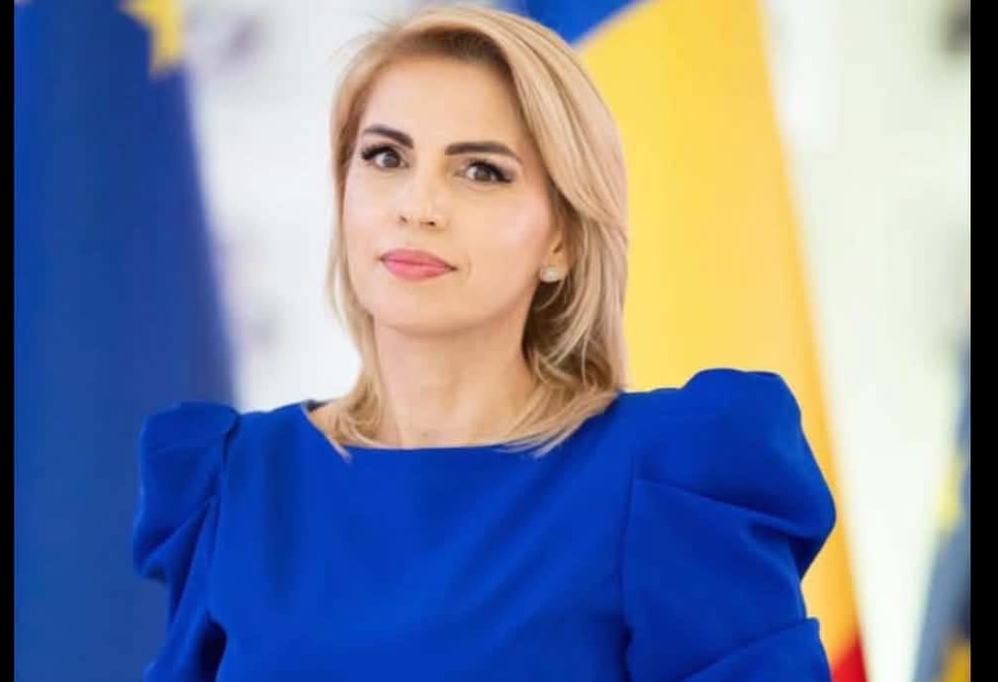 Screenshot 2026-10-23 162548 - Jurnalul Olteniei – Știri din Craiova și Oltenia Professional woman in a blue dress gazing slightly to the left, with blurred flags in the background indicating a formal event or press setting.