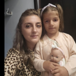 Adult woman and young girl pose for a selfie, with the girl holding a baby doll in a pale pink cardigan.