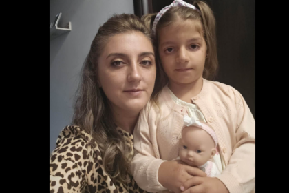 Adult woman and young girl pose for a selfie, with the girl holding a baby doll in a pale pink cardigan.