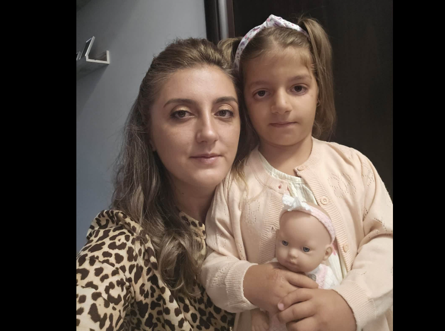 Adult woman and young girl pose for a selfie, with the girl holding a baby doll in a pale pink cardigan.