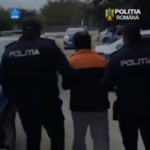 Two Romanian police officers escort a man in an orange jacket on a street. The officers wear dark uniforms with POLITIA printed on the back (scene outdoors).