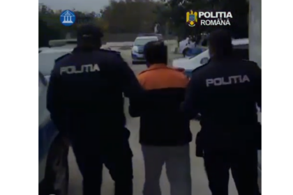 Two Romanian police officers escort a man in an orange jacket on a street. The officers wear dark uniforms with POLITIA printed on the back (scene outdoors).