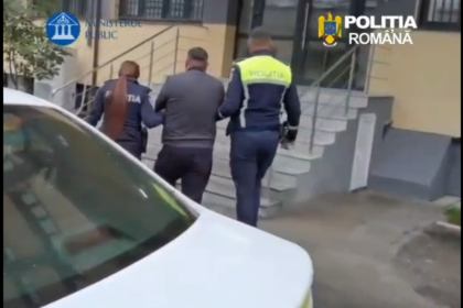 Two Romanian police officers escort a man up exterior stairs toward a building, with Ministerul Public and Politia Romana logos visible.