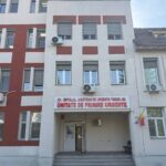 Facade of a hospital building with a red-and-beige pattern; sign above entrance reads 'Unitate de Primire Urgente' (Emergency Department) in Romanian.