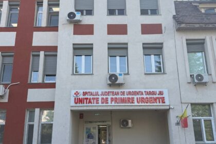 Facade of a hospital building with a red-and-beige pattern; sign above entrance reads 'Unitate de Primire Urgente' (Emergency Department) in Romanian.