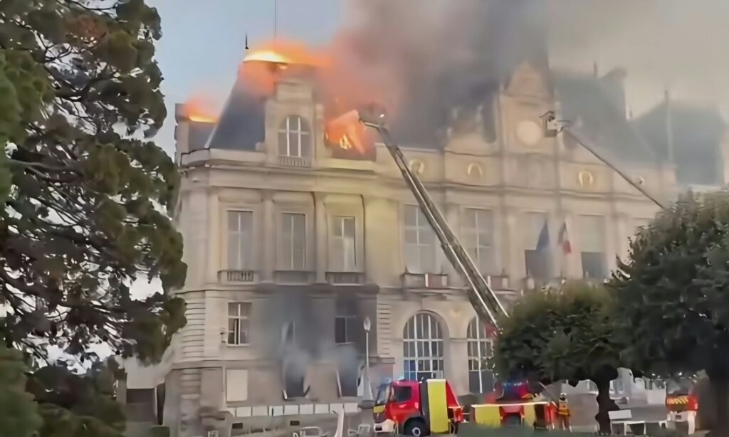 Rooftop of a beige multi-story building ablaze with flames and smoke; a fire truck with an aerial ladder on scene.