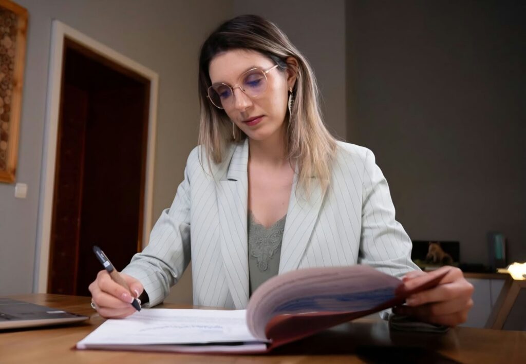 Woman with light hair and round glasses sits at a desk, signing a document with a pen in a professional setting.