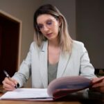 Woman with light hair and round glasses sits at a desk, signing a document with a pen in a professional setting.