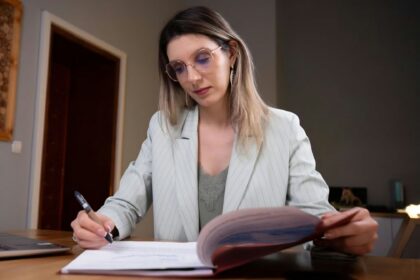 Woman with light hair and round glasses sits at a desk, signing a document with a pen in a professional setting.