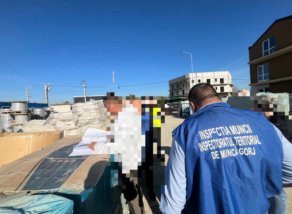 Blue-vested official from the Gorj Territorial Labour Inspectorate reviewing documents at a construction site with pallets and building materials around him while a group of people looks on.