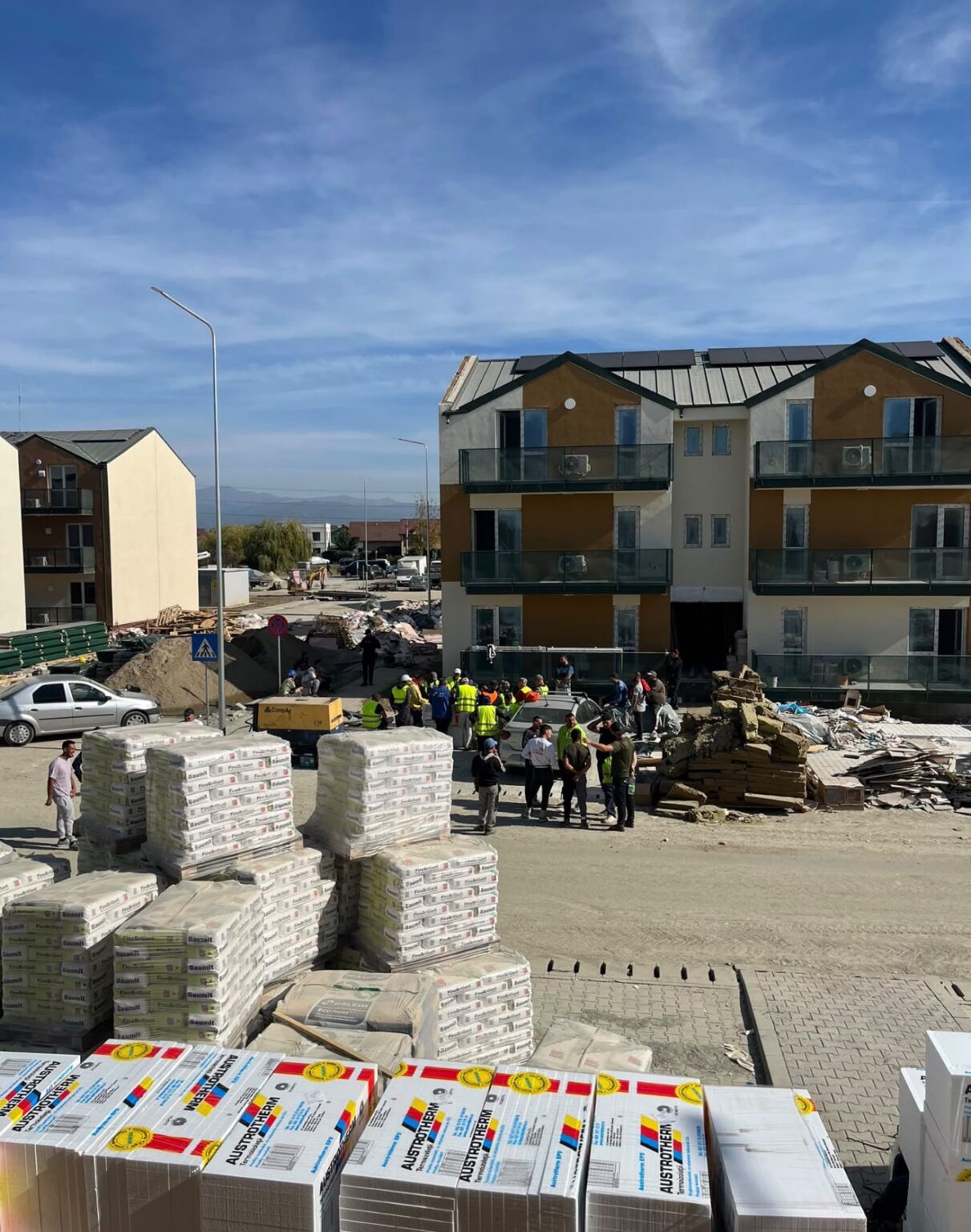 Construction site in a residential area with workers and stacked building materials in front of a three-story building complex.