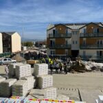 Construction site in a residential area with workers and stacked building materials in front of a three-story building complex.