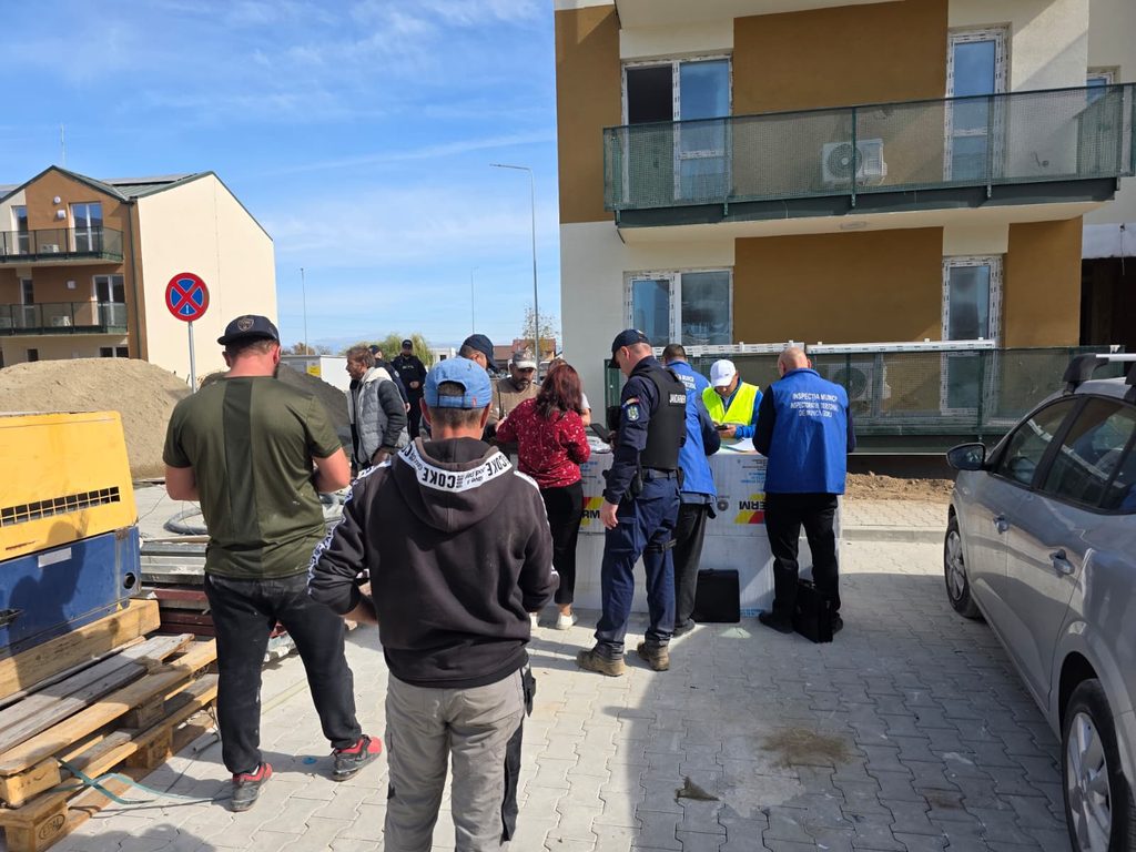 Group of people, including inspectors in blue vests, gather around a temporary barrier at a residential construction site with pallets and sand nearby.