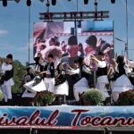 Group of dancers in traditional black-and-white outfits performing a folk dance on an outdoor festival stage with a banner reading Festivalul Tocanului.