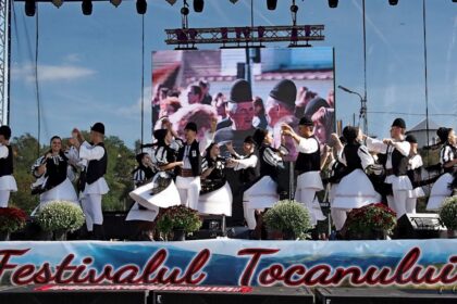 Group of dancers in traditional black-and-white outfits performing a folk dance on an outdoor festival stage with a banner reading Festivalul Tocanului.
