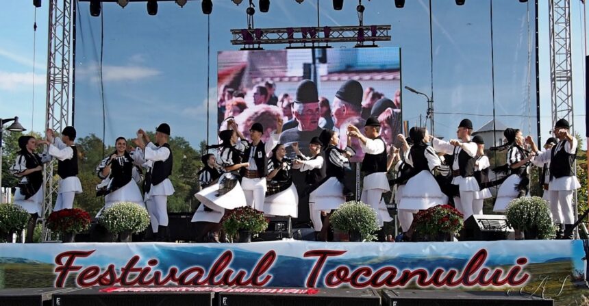Group of dancers in traditional black-and-white outfits performing a folk dance on an outdoor festival stage with a banner reading Festivalul Tocanului.