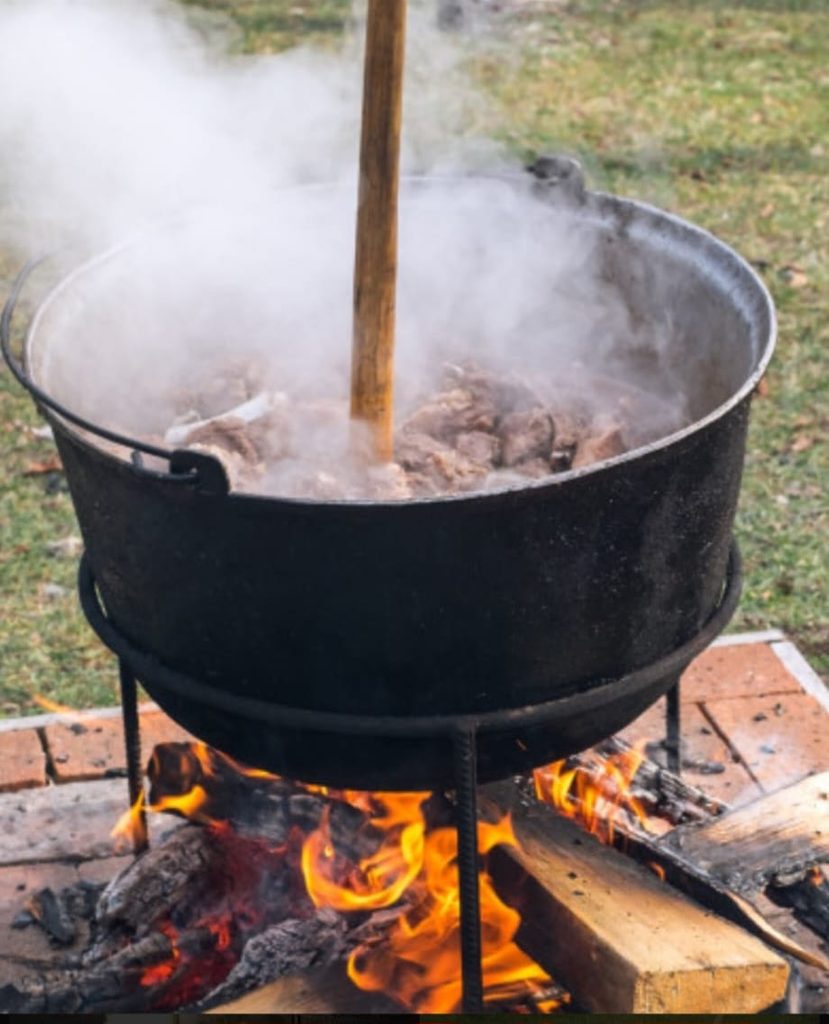 Large black cast-iron cauldron on a tripod over a wood fire, steam rising as meat cooks outdoors.
