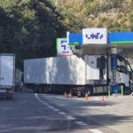 Gas station fueling area with two large white trucks and orange cones, forested hillside in the background.