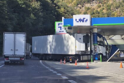 Gas station fueling area with two large white trucks and orange cones, forested hillside in the background.