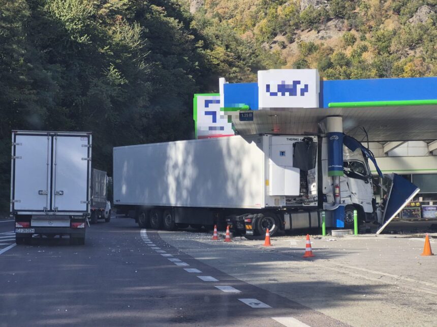 Gas station fueling area with two large white trucks and orange cones, forested hillside in the background.