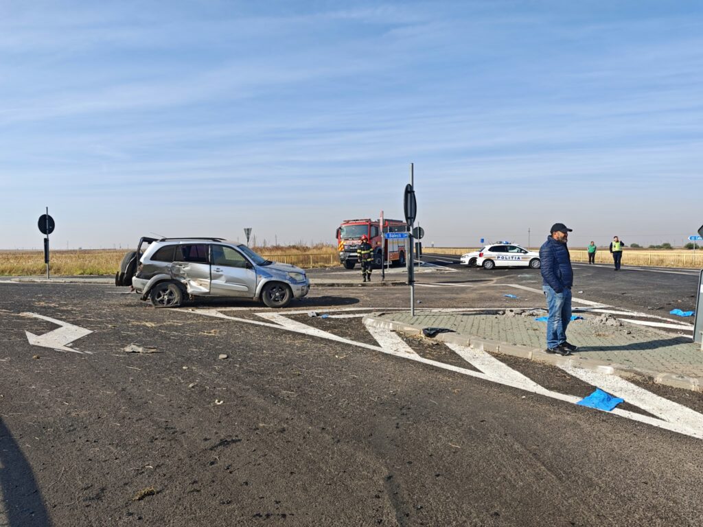 Silver SUV with significant front-end damage sits across a road, emergency responders and a fire truck nearby under a clear blue sky, suggesting a traffic accident.