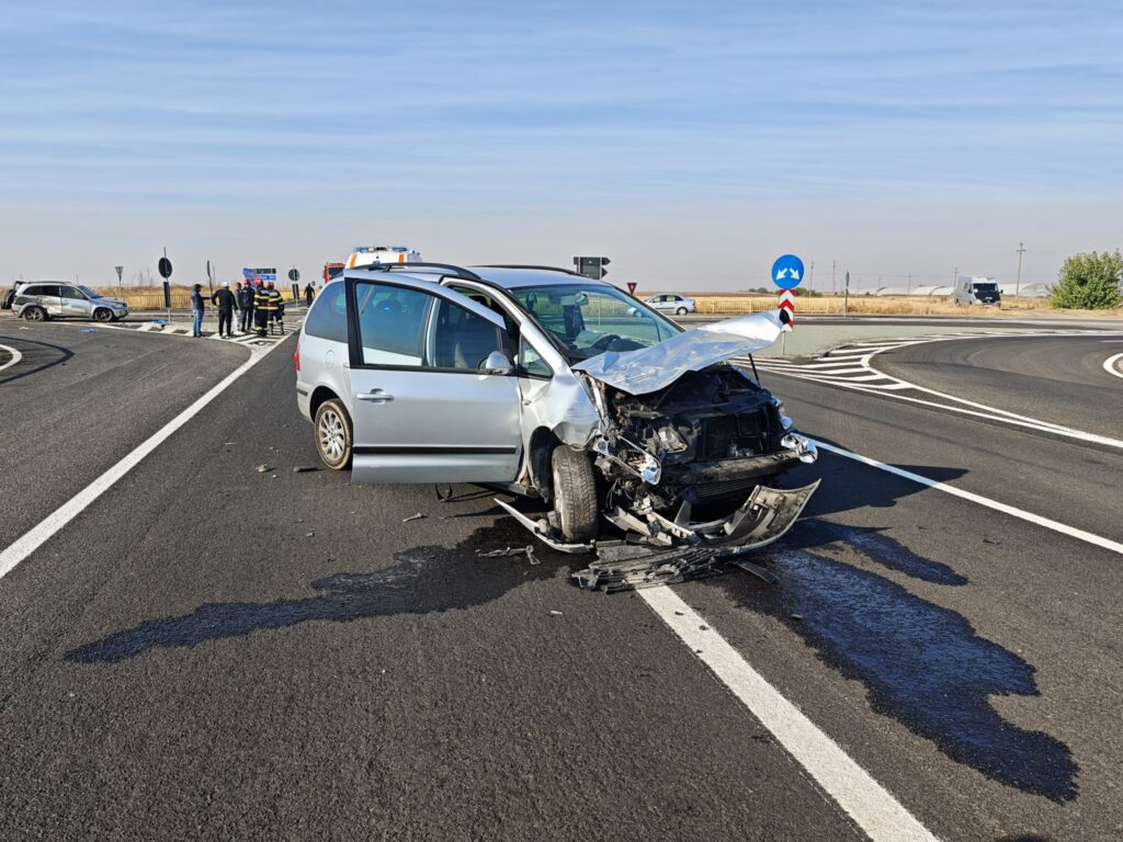 Silver minivan with heavy front-end damage at a roundabout after a crash; emergency responders and other vehicles in the background.