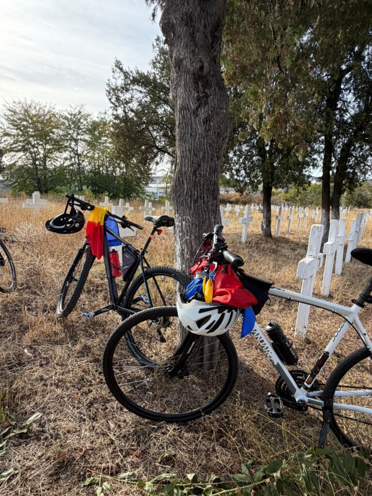 Two bicycles leaning against a tree in a graveyard with white crosses and dry grass under a cloudy sky.