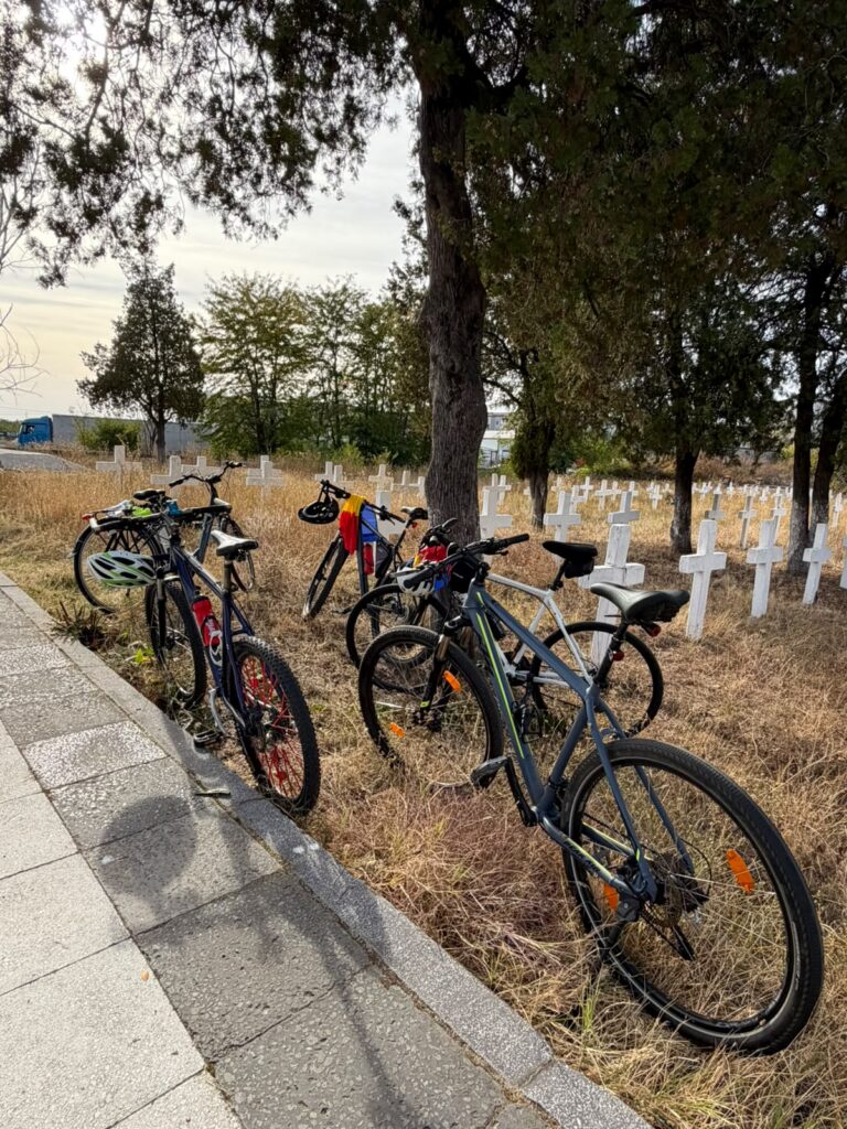 Bicycles parked along a paved path beside a dry grass cemetery with white crosses and trees in the background.