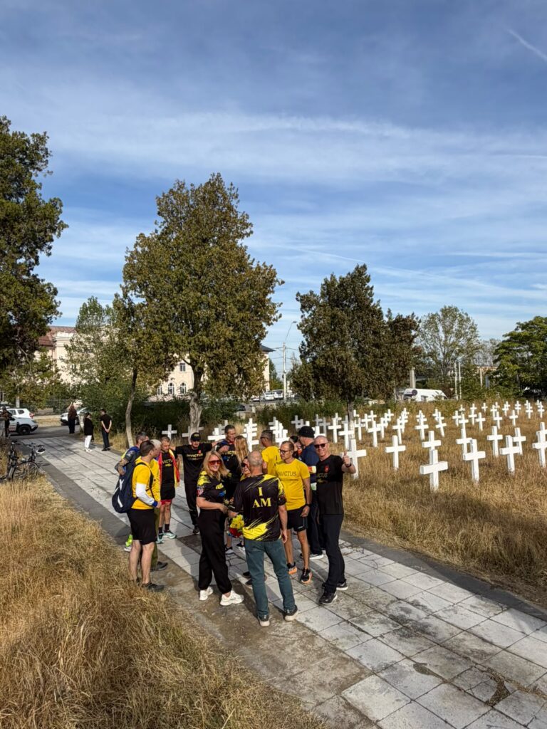 Group of people in yellow and black shirts gather on a stone path beside a field of white crosses, likely a memorial, with trees and a blue sky in the background.