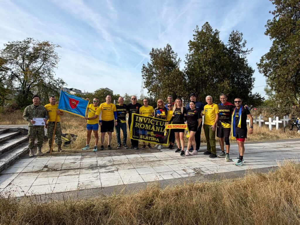 Group of people wearing yellow and black Invictus Romania shirts posing outdoors with banners; a man in uniform holds a certificate in the foreground.