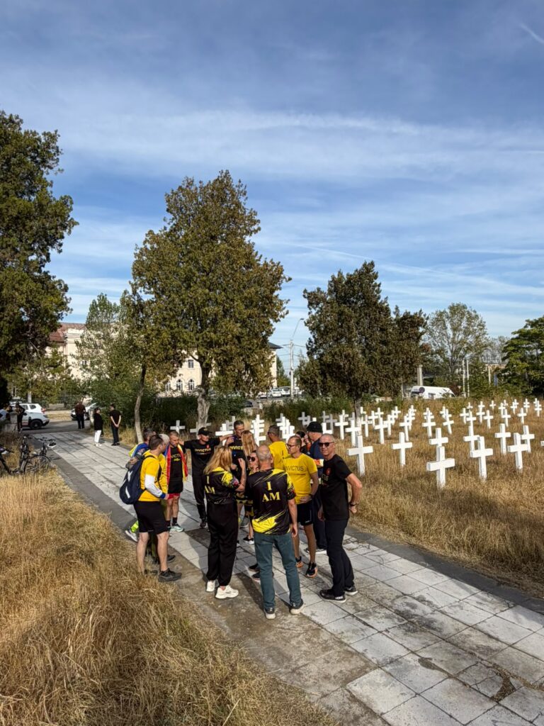 Group of people in yellow and black shirts stand on a paved path beside a field of white crosses in a cemetery under a blue sky.