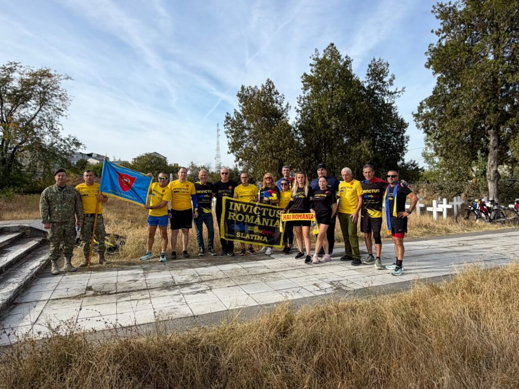 Group of about 18 people posing outdoors on a paved plaza, several wearing yellow Invictus shirts, holding a blue flag with a red shield and a banner reading'INVICTUS ROMANIA SLATINA'; soldiers in uniform stand to the left; others hold a yellow and black Invictus banner and a 'HAI ROMANIA' sign, with trees and clear blue sky in the background.