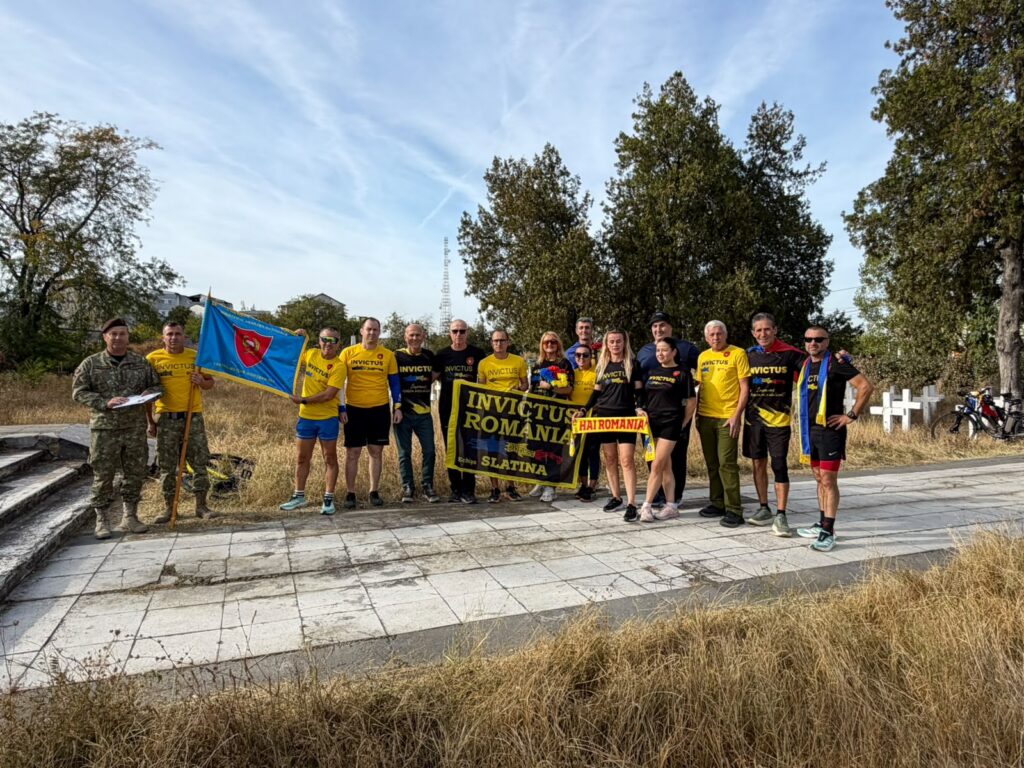 Group of people in yellow Invictus Romania shirts pose with flags and banners outdoors on a sunny day.