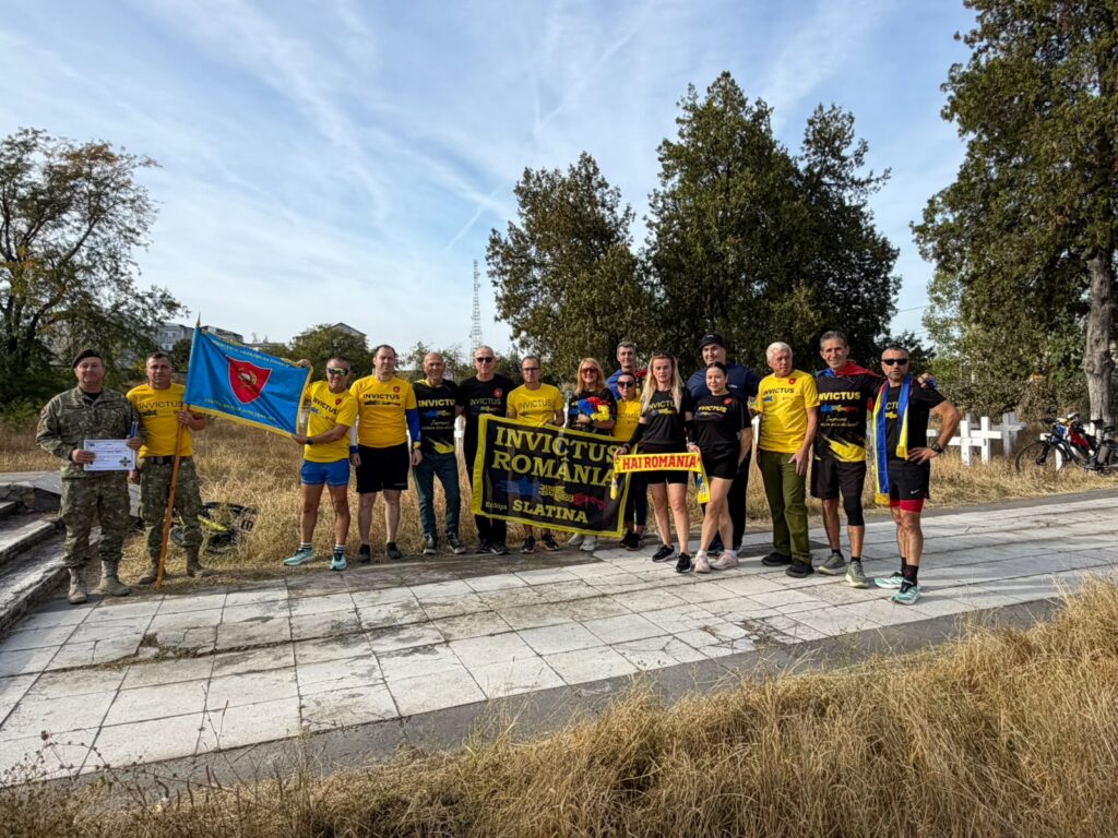 Group of Invictus Romania athletes wearing yellow shirts, posing outdoors with banners and a blue flag on a sunny day.