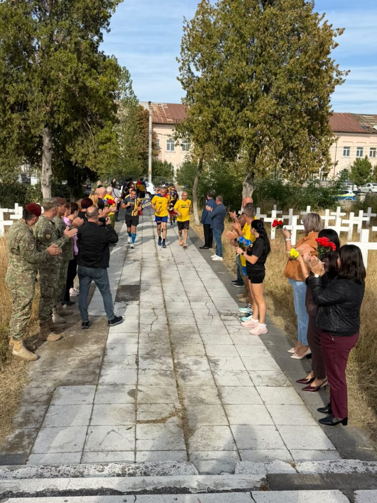Runners in yellow shirts finish a race as crowds clap along a stone path flanked by trees and civilians holding flowers.