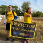 Two people wearing yellow Invictus shirts stand in a grassy area, holding a black and yellow Invictus Team Romania banner in front of a cemetery with white crosses.