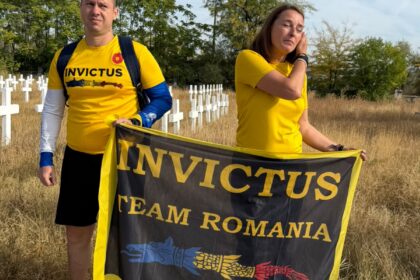 Two people wearing yellow Invictus shirts stand in a grassy area, holding a black and yellow Invictus Team Romania banner in front of a cemetery with white crosses.